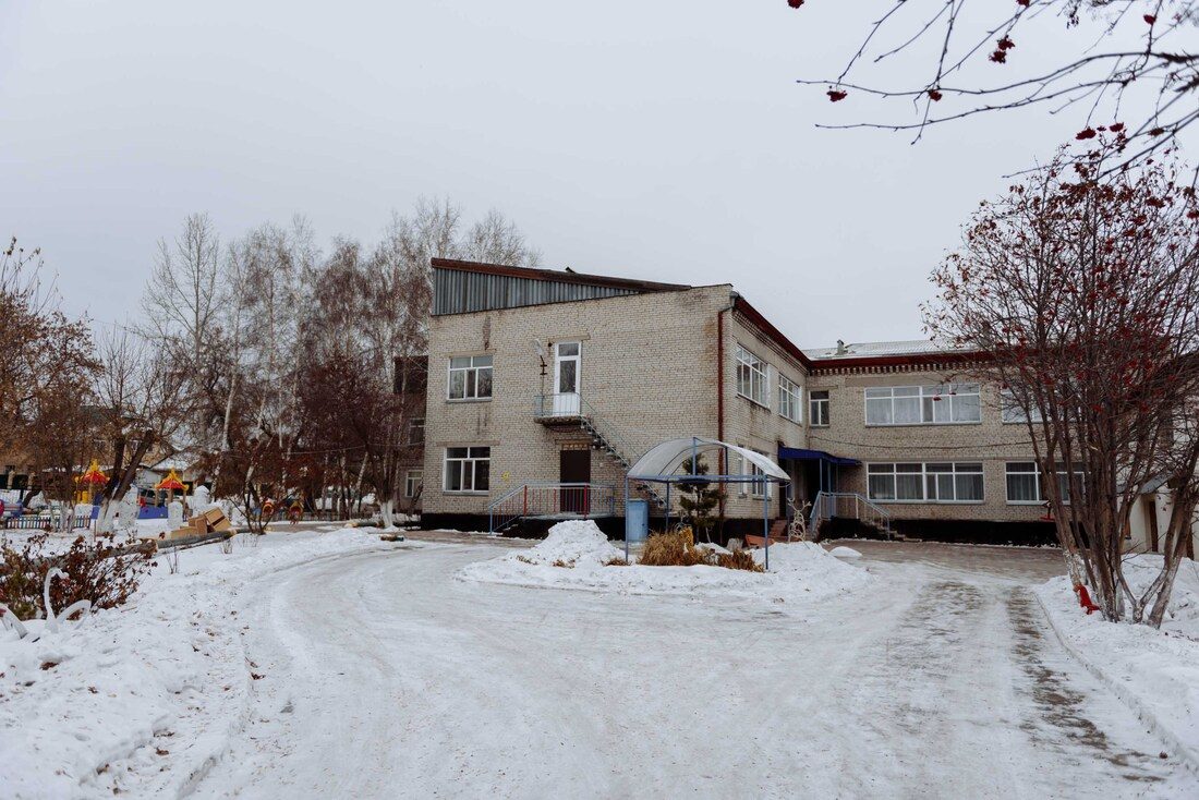 The two-storey pale brick Shchuchinsk Babyhouse in winter, with a snow-covered driveway and a playground to one side