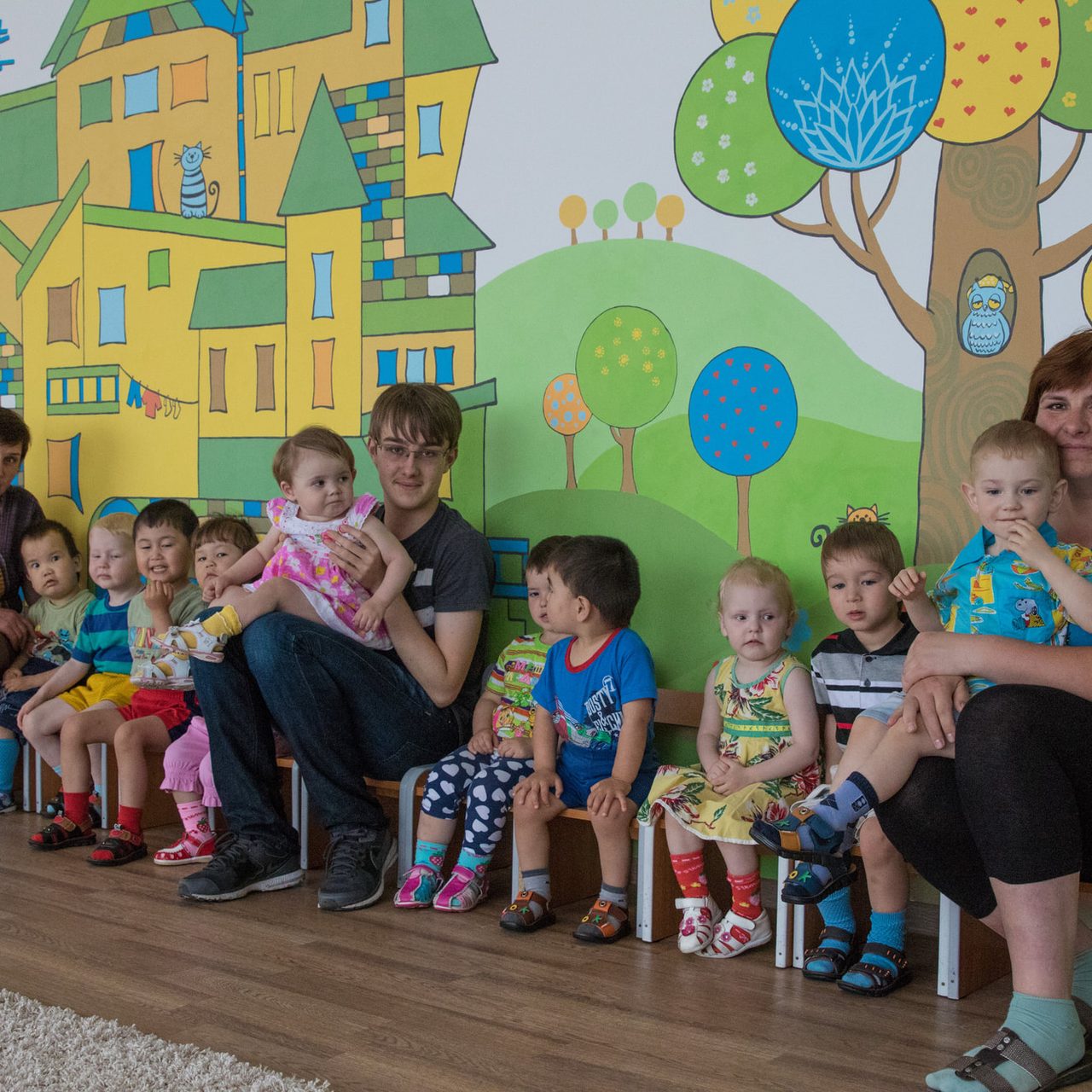 Two caregivers and a teenage volunteer sitting on a bench with a dozen toddlers in front of a brightly painted mural at the Shchuchinsk Babyhouse