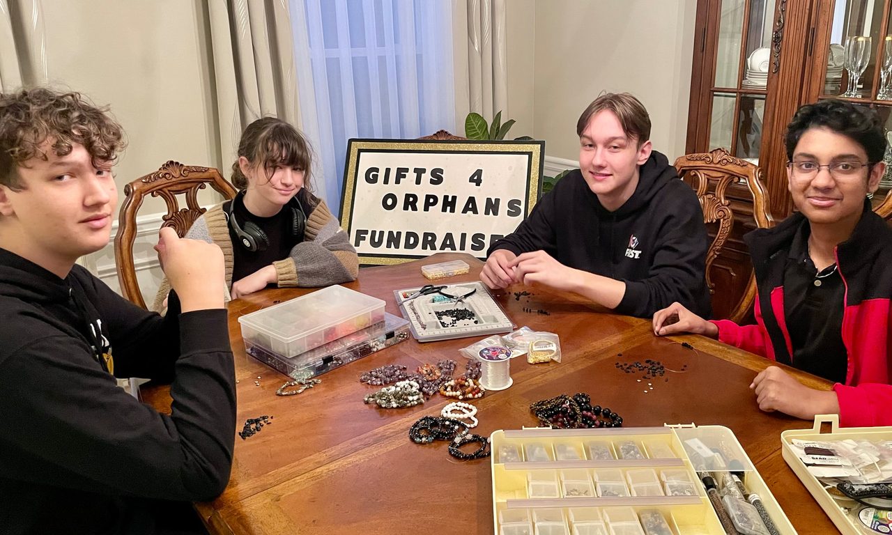 Four student volunteers making beaded jewelry around a dining table behind a hand-lettered “Gifts 4 Orphans Fundraiser” sign