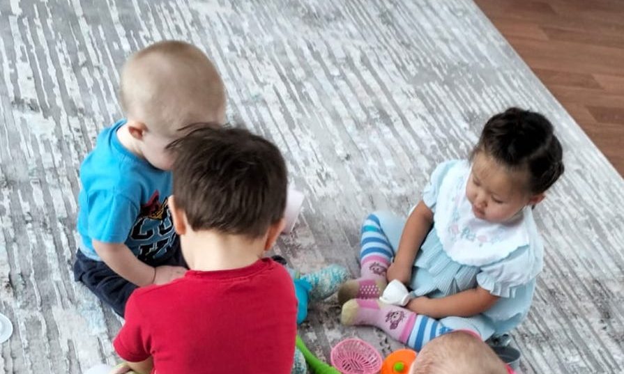 Four toddlers sitting and playing with toys on a new grey patterned carpet