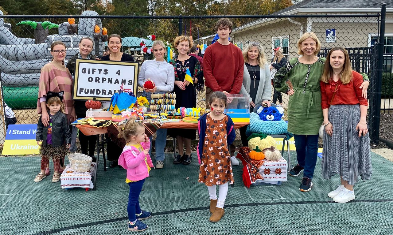 A group of volunteers and children standing behind a fundraising table of handmade crafts, with Ukrainian flags and an “I Stand With Ukraine” sign