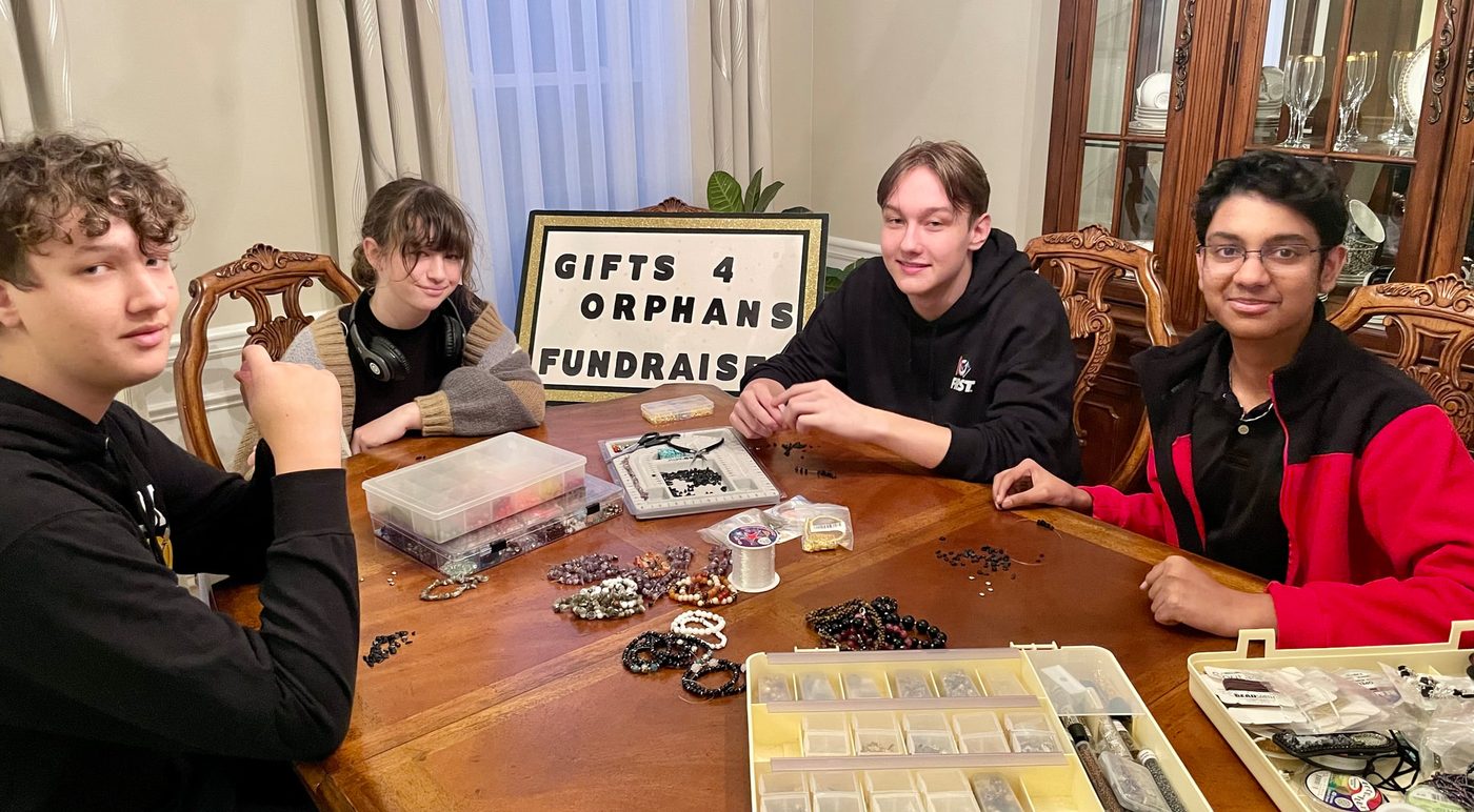 Four student volunteers making beaded jewelry around a dining table behind a "Gifts 4 Orphans Fundraiser" sign