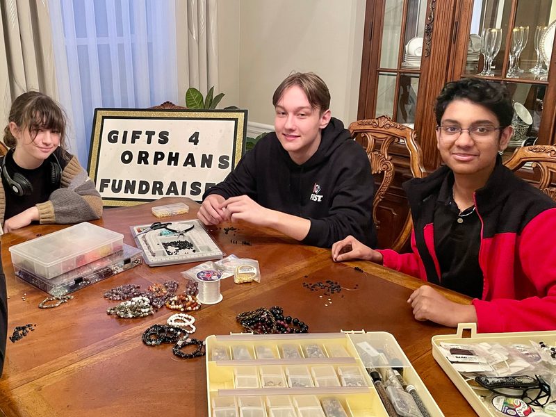 Four student volunteers making beaded jewelry around a dining table behind a "Gifts 4 Orphans Fundraiser" sign