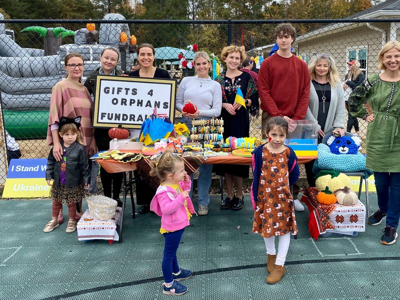 Volunteers and children behind a fundraising table with handmade crafts, Ukrainian flags and an "I Stand With Ukraine" sign